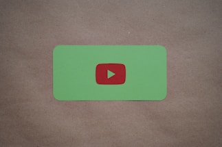 A green card with rounded corners features a prominent red play button symbol in the center, set against a plain brown textured background.