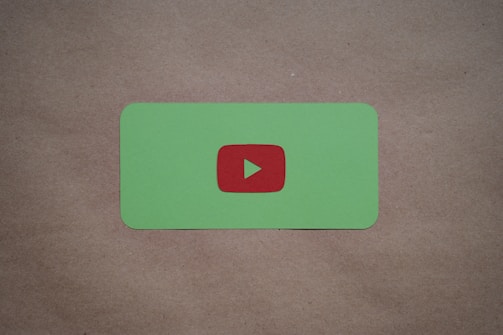 A green card with rounded corners features a prominent red play button symbol in the center, set against a plain brown textured background.