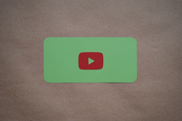 A green card with rounded corners features a prominent red play button symbol in the center, set against a plain brown textured background.