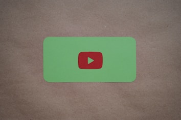 A green card with rounded corners features a prominent red play button symbol in the center, set against a plain brown textured background.
