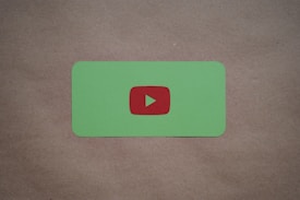 A green card with rounded corners features a prominent red play button symbol in the center, set against a plain brown textured background.