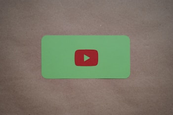A green card with rounded corners features a prominent red play button symbol in the center, set against a plain brown textured background.
