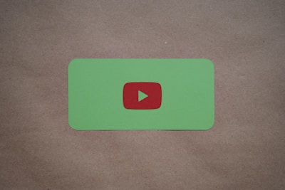 A green card with rounded corners features a prominent red play button symbol in the center, set against a plain brown textured background.