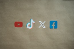 Four social media platform logos are arranged on a brown background. From left to right, the logos are for YouTube in red, TikTok in a combination of red, blue, and white, X in white, and Facebook in blue.