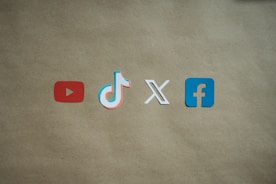 Four social media platform logos are arranged on a brown background. From left to right, the logos are for YouTube in red, TikTok in a combination of red, blue, and white, X in white, and Facebook in blue.