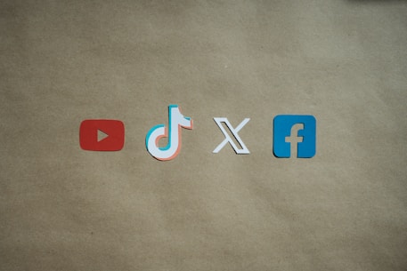 Four social media platform logos are arranged on a brown background. From left to right, the logos are for YouTube in red, TikTok in a combination of red, blue, and white, X in white, and Facebook in blue.