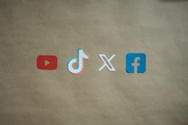 Four social media platform logos are arranged on a brown background. From left to right, the logos are for YouTube in red, TikTok in a combination of red, blue, and white, X in white, and Facebook in blue.