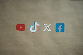 Four social media platform logos are arranged on a brown background. From left to right, the logos are for YouTube in red, TikTok in a combination of red, blue, and white, X in white, and Facebook in blue.