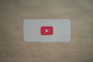 A white rectangular card with rounded corners is placed on a brown surface. In the center of the card, there is a red play button icon resembling the YouTube logo.