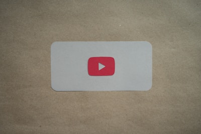 A white rectangular card with rounded corners is placed on a brown surface. In the center of the card, there is a red play button icon resembling the YouTube logo.