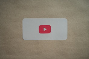 A white rectangular card with rounded corners is placed on a brown surface. In the center of the card, there is a red play button icon resembling the YouTube logo.