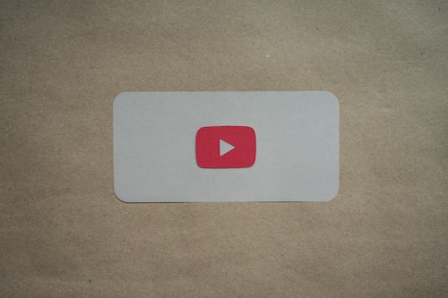 A white rectangular card with rounded corners is placed on a brown surface. In the center of the card, there is a red play button icon resembling the YouTube logo.