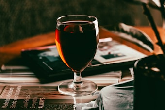 A glass of red wine sits on top of several magazines on a table. Soft lighting creates a warm ambiance, with shadows and reflections visible on the wine glass. In the background, a blurred image of an open magazine and a plant are slightly visible.