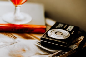A close-up of a vintage camera lying on a table with a blurred magazine visible beneath it. Beside the camera, a book with a bright orange cover can be seen, and a glass containing a red liquid is partially visible in the background.