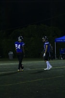 Two football players in blue jerseys are standing on a dimly lit football field. One player is wearing the number 24. There is a blue tent with text and some other individuals in the background.