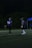 Two football players in blue jerseys are standing on a dimly lit football field. One player is wearing the number 24. There is a blue tent with text and some other individuals in the background.
