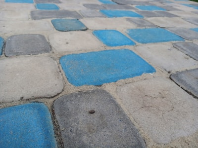 A pattern of square paving stones in a repeating arrangement of blue and gray hues. The stones have rough textures and are set in a slightly worn surface, appearing to form a path or patio.