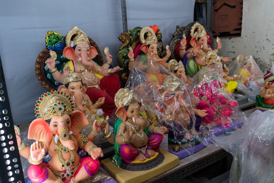 The image features a collection of intricately designed statues of the Hindu deity Ganesha, each adorned with colorful clothing, ornate jewelry, and elaborate golden crowns. Some statues are wrapped in protective plastic. They are arranged in rows on display for sale, showcasing various poses and vibrant colors.