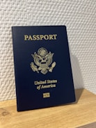 A United States passport stands upright on a wooden surface against a textured white wall. The passport is dark blue with gold embossed text and emblem on the cover.