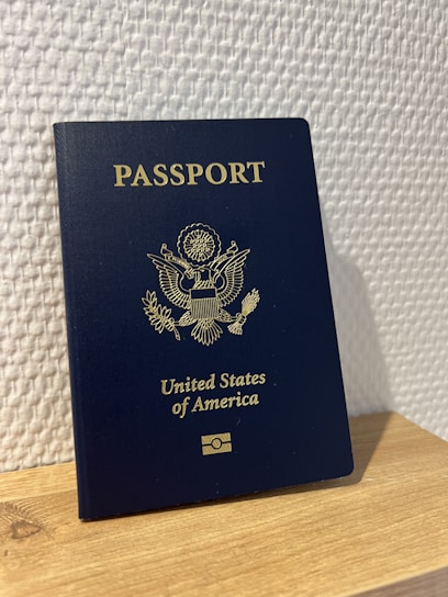 A United States passport stands upright on a wooden surface against a textured white wall. The passport is dark blue with gold embossed text and emblem on the cover.