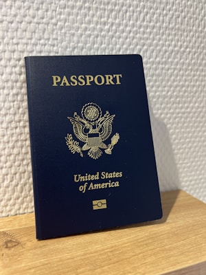 A United States passport stands upright on a wooden surface against a textured white wall. The passport is dark blue with gold embossed text and emblem on the cover.