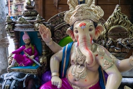 A large statue of Ganesha, the Hindu deity with the head of an elephant, adorned with an ornate crown and jewelry, sits beside a smaller seated figure dressed in pink with an elaborate turban. The backdrop includes intricate engravings and decorative elements.