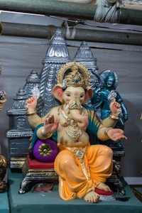 The image displays a statue of the Hindu deity Ganesha, adorned with elaborate golden jewelry and a royal headdress. He is depicted seated on a throne, with a serene expression and multiple hands holding various attributes such as a flower, an axe, and a modak (sweet). Behind the statue are smaller figures and ornate temple-like structures.