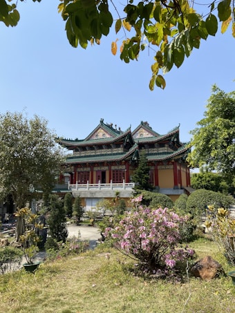 A traditional Asian temple with ornate, green-tiled roofs sits in the background, surrounded by lush gardens and trees. In the foreground, pink flowers bloom amidst greenery, while the sky overhead is clear and blue with some leaves framing the top of the image.