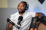 A person wearing headphones is seated on a couch, speaking into a microphone. The individual is dressed in a white shirt and appears to be relaxed. There is audio equipment visible, hinting at a podcast or radio show setting.