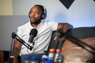 A person wearing headphones is speaking into a microphone while sitting on a sofa. Various audio equipment, including a microphone stand, is visible. Several water bottles are placed on the surface near the person, with some foam padding on the wall in the background.