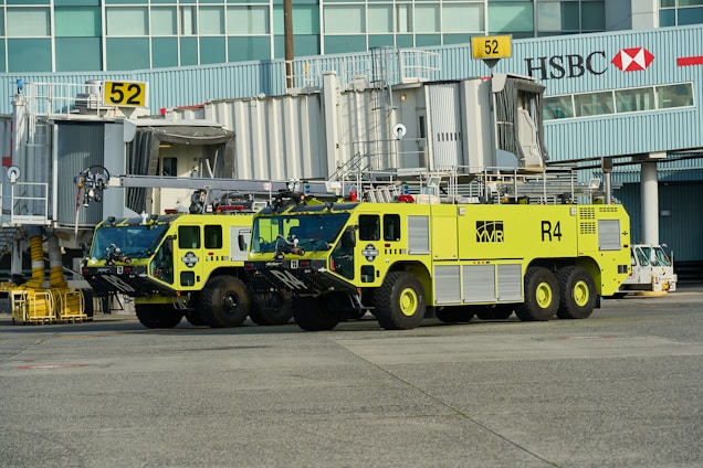 Two large, lime-green airport fire trucks are parked in front of gate 52 at an airport terminal. The vehicles are equipped with fire fighting equipment and have 'R4' markings. The background includes passenger boarding bridges, airport signage and the HSBC logo on a building facade.