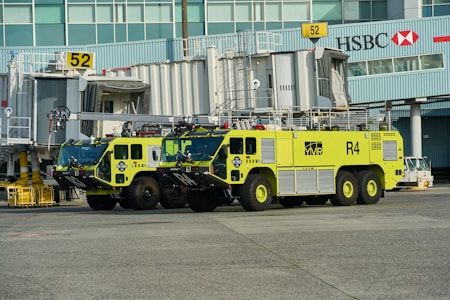 Two large, lime-green airport fire trucks are parked in front of gate 52 at an airport terminal. The vehicles are equipped with fire fighting equipment and have 'R4' markings. The background includes passenger boarding bridges, airport signage and the HSBC logo on a building facade.
