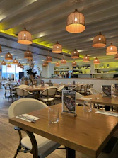 A well-lit restaurant interior with wooden tables and chairs neatly arranged. The ceiling has hanging wicker lamps, casting a warm glow. Walls are adorned with shelves holding bottles, and there is a bar area in the background. Menus titled 'Meat Platter' are placed on the tables.