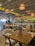 A well-lit restaurant interior with wooden tables and chairs neatly arranged. The ceiling has hanging wicker lamps, casting a warm glow. Walls are adorned with shelves holding bottles, and there is a bar area in the background. Menus titled 'Meat Platter' are placed on the tables.