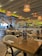A well-lit restaurant interior with wooden tables and chairs neatly arranged. The ceiling has hanging wicker lamps, casting a warm glow. Walls are adorned with shelves holding bottles, and there is a bar area in the background. Menus titled 'Meat Platter' are placed on the tables.