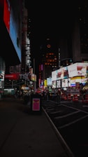 A nighttime urban cityscape featuring a bustling street filled with large, brightly lit billboards and advertisements. Construction cones and signs are visible near the lower right, with a few people walking along the sidewalk. The area appears to be busy despite the late hour, emphasizing the vibrant and lively atmosphere.
