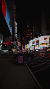 A nighttime urban cityscape featuring a bustling street filled with large, brightly lit billboards and advertisements. Construction cones and signs are visible near the lower right, with a few people walking along the sidewalk. The area appears to be busy despite the late hour, emphasizing the vibrant and lively atmosphere.