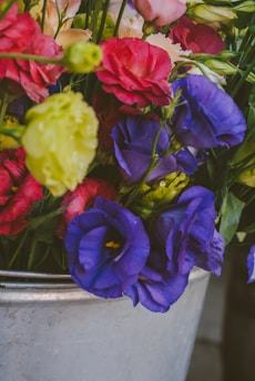 A variety of colorful flowers including purple, red, pink, and yellow blooms are arranged in a metal bucket. The vibrant petals and abundant greenery create a lively and fresh appearance.