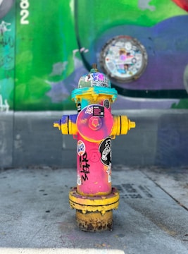 A brightly colored fire hydrant covered in various stickers is placed on a concrete pavement. The hydrant is predominantly pink with yellow accents and features eclectic designs. A graffiti-covered wall serves as a vibrant backdrop with shades of green and purple.