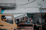 A small roadside shop with a variety of snacks and goods displayed. A man is sitting inside a parked yellow truck, looking at a mobile phone. The shop has a tarp roof, and various items are hanging, including packets and bottles. The walls are worn and there are posters affixed to them.
