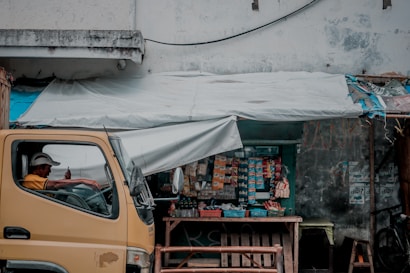 A small roadside shop with a variety of snacks and goods displayed. A man is sitting inside a parked yellow truck, looking at a mobile phone. The shop has a tarp roof, and various items are hanging, including packets and bottles. The walls are worn and there are posters affixed to them.