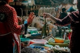 A bustling market scene with two individuals engaged in a transaction. Various fresh produce, such as vegetables and fruits, are displayed on stalls. The atmosphere is vibrant, with different textures and colors from the goods adding to the lively ambiance.