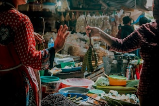 A bustling market scene with two individuals engaged in a transaction. Various fresh produce, such as vegetables and fruits, are displayed on stalls. The atmosphere is vibrant, with different textures and colors from the goods adding to the lively ambiance.