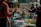 A bustling market scene with two individuals engaged in a transaction. Various fresh produce, such as vegetables and fruits, are displayed on stalls. The atmosphere is vibrant, with different textures and colors from the goods adding to the lively ambiance.