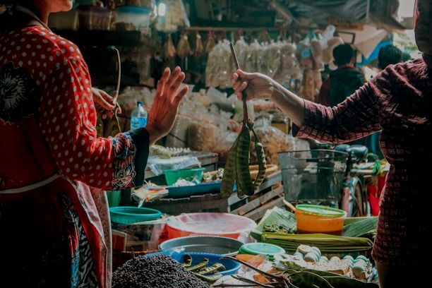 A bustling market scene with two individuals engaged in a transaction. Various fresh produce, such as vegetables and fruits, are displayed on stalls. The atmosphere is vibrant, with different textures and colors from the goods adding to the lively ambiance.