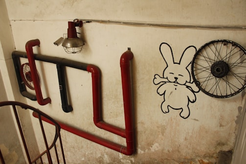 A wall art installation featuring red and black pipes mounted on a textured beige wall. A vintage industrial lamp is attached above the pipes. To the right, a simplistic drawing of a rabbit holding a bone is painted on the wall. A bicycle wheel without a tire is mounted next to the drawing, adding an eclectic touch.