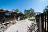 An outdoor patio area adjacent to a house with large windows. The patio is paved with concrete and has a section with decorative stones and plants. There is a metal fence running along the right side, and a plastic chair is placed near the fence. Trees are visible in the background against a clear blue sky.