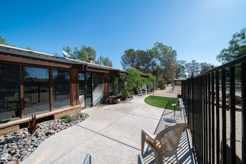 An outdoor patio area adjacent to a house with large windows. The patio is paved with concrete and has a section with decorative stones and plants. There is a metal fence running along the right side, and a plastic chair is placed near the fence. Trees are visible in the background against a clear blue sky.