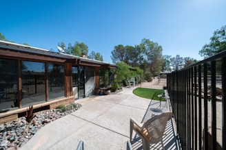 An outdoor patio area adjacent to a house with large windows. The patio is paved with concrete and has a section with decorative stones and plants. There is a metal fence running along the right side, and a plastic chair is placed near the fence. Trees are visible in the background against a clear blue sky.