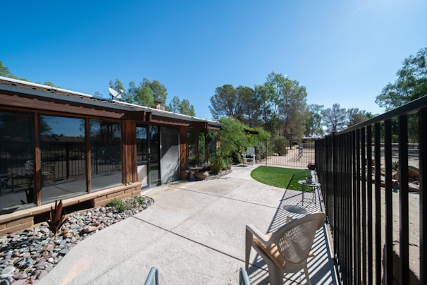 An outdoor patio area adjacent to a house with large windows. The patio is paved with concrete and has a section with decorative stones and plants. There is a metal fence running along the right side, and a plastic chair is placed near the fence. Trees are visible in the background against a clear blue sky.